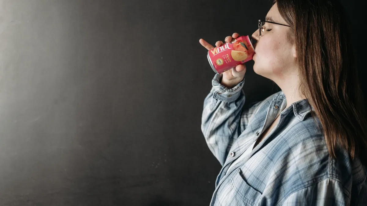 A woman in a casual shirt drinks from a canned beverage against a dark background.