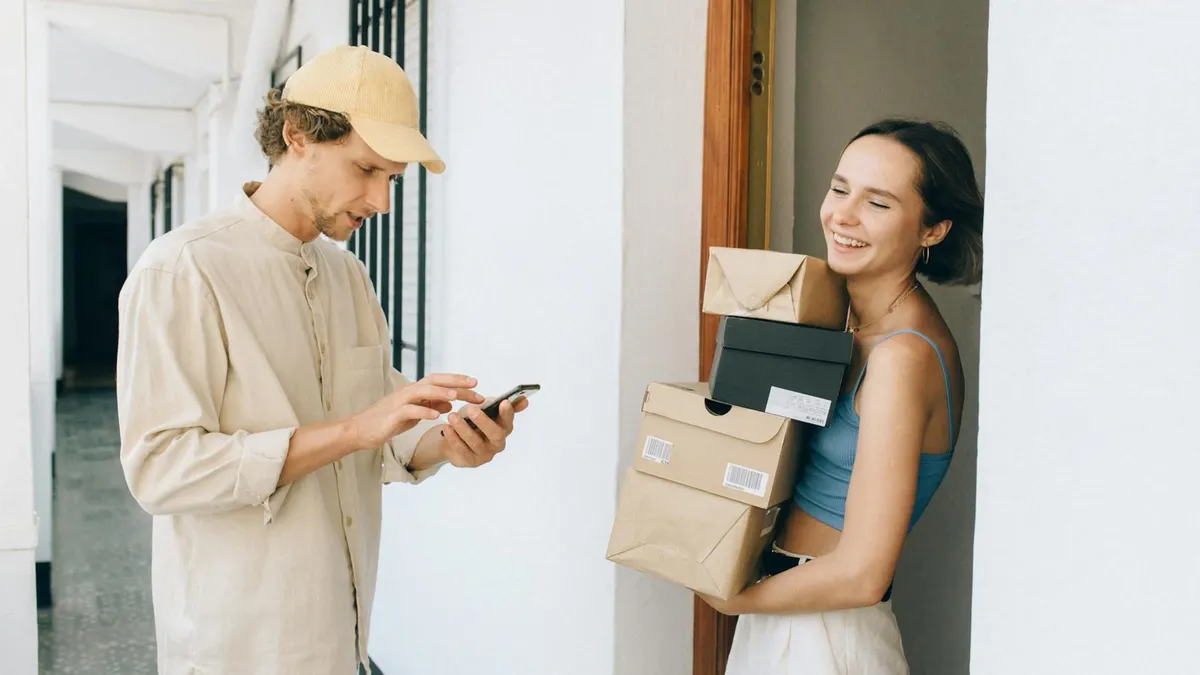 A cheerful woman receives a delivery from a person in a hallway, using a smartphone for online payment.