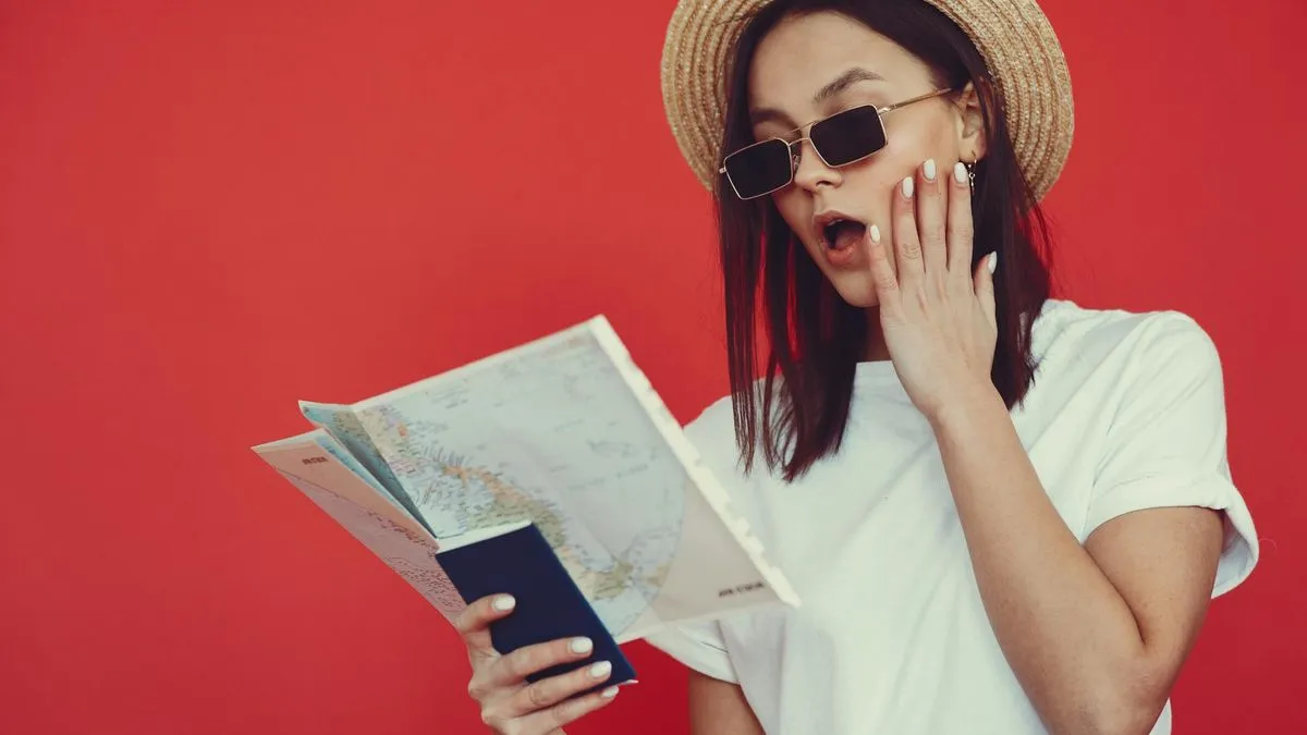 A surprised woman in a straw hat and sunglasses holds a map and passport against a red background.