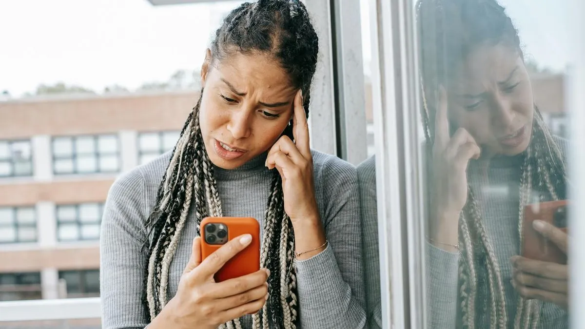 A woman with braided hair looks frustrated while holding and looking at her phone.