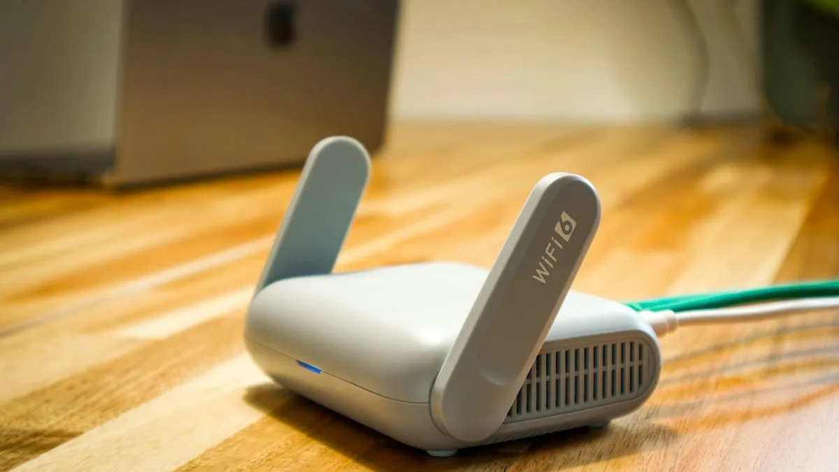 A white Wi-Fi 6 router with two antennas stands on a wooden table, connected by a green cable.