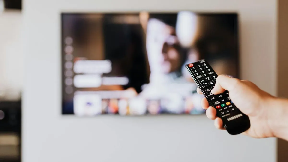 A hand holding a TV remote control, with a blurred television screen in the background.