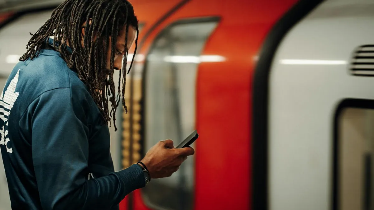 Young man with dreadlocks using smartphone on London Underground platform, waiting for train.