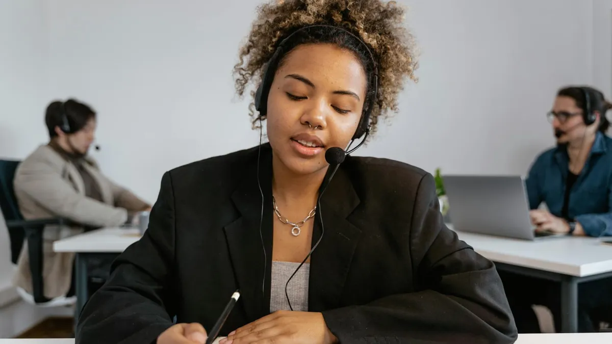 African American woman with headset working in an office with colleagues, using a pen for note-taking.