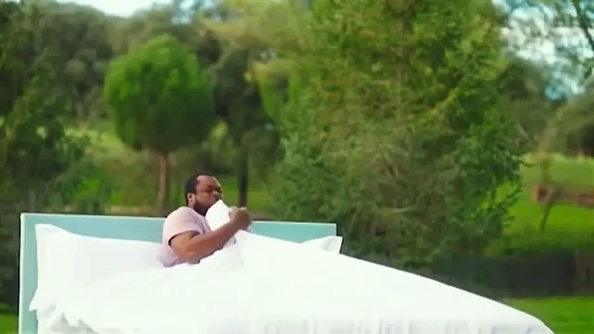 A man with a beard and short hair sits up in a bed with white sheets outdoors in a green field.