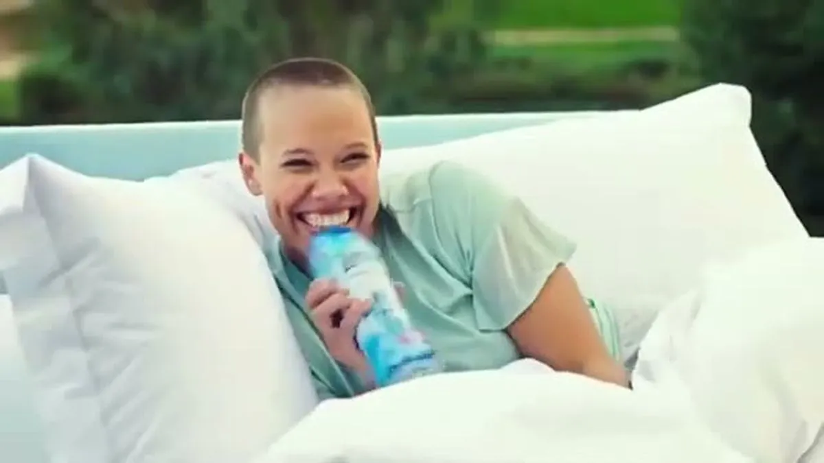 A woman with a shaved head laughs, holding a blue Lenor bottle while lying in a bed outdoors.