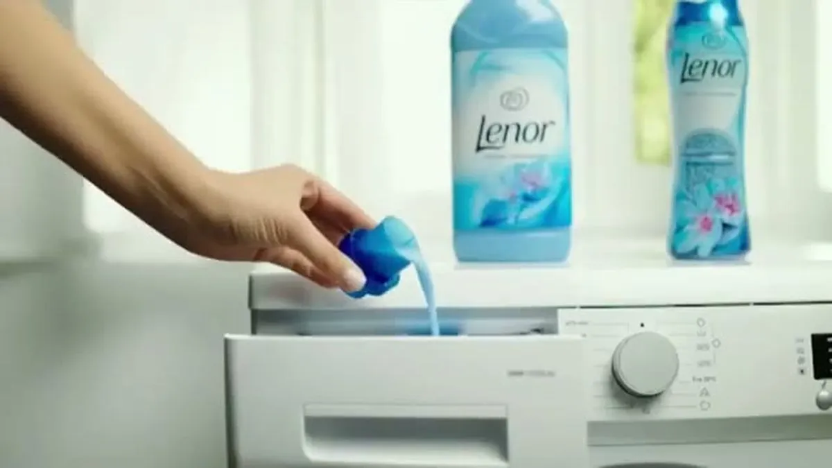 A hand pours blue Lenor fabric conditioner from a measuring cup into a washing machine drawer.
