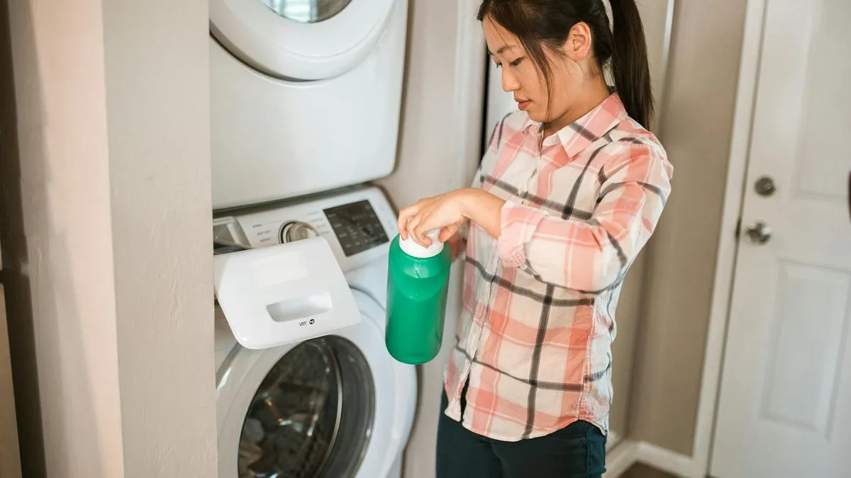A woman in a plaid shirt pours liquid detergent into a washing machine.