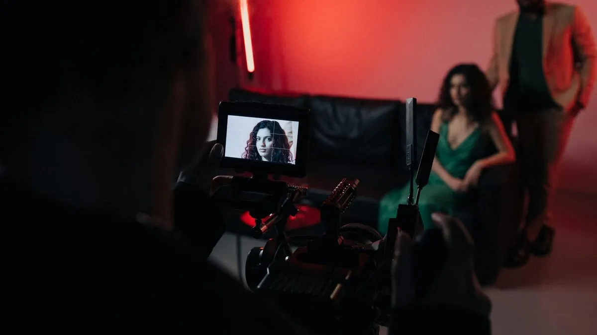 A camera operator films a woman in a green dress on a black couch, with a man standing behind.