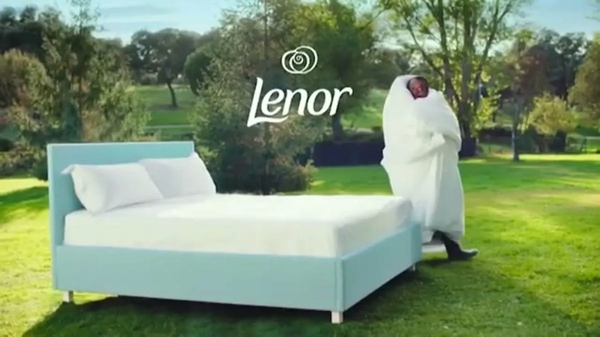 A wide shot of a bed in a grassy field with a person covered in white fabric standing beside it.