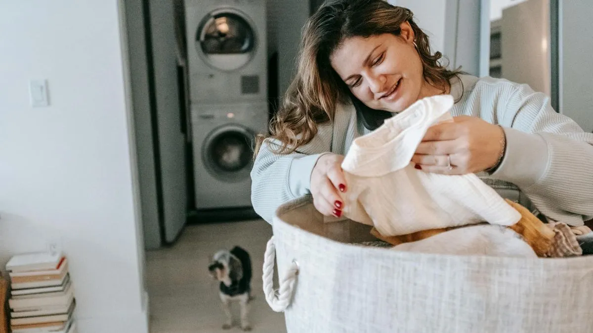 A smiling woman in a light sweater holds a white cloth over a laundry basket.